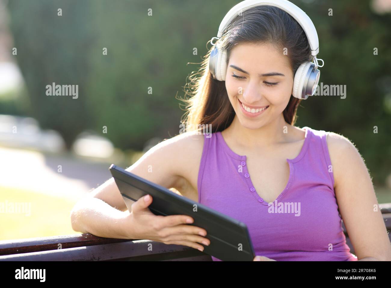 Donna felice che guarda i contenuti multimediali sul tablet con le cuffie sedute in un parco Foto Stock