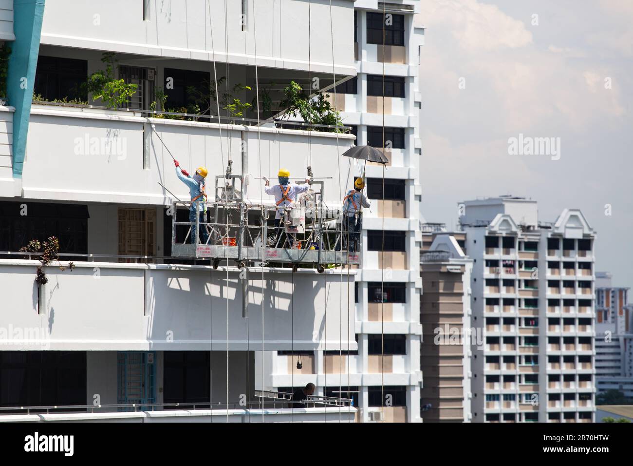 Lavoratori che utilizzano un sollevatore industriale sospeso a mezz'aria che eseguono lavori di verniciatura per un esterno di un edificio sotto il sole caldo. Un'occupazione a rischio molto elevato. Foto Stock