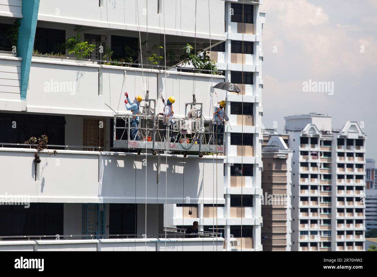 Lavoratori che utilizzano un sollevatore industriale sospeso a mezz'aria che eseguono lavori di verniciatura per un esterno di un edificio sotto il sole caldo. Un'occupazione a rischio molto elevato. Foto Stock