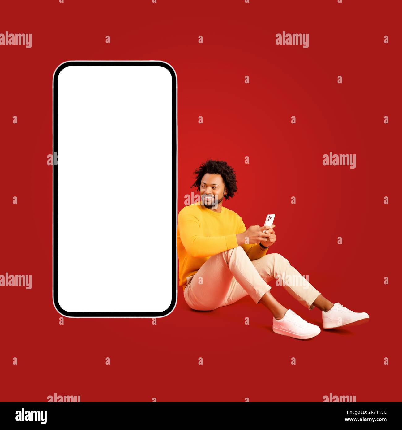 Bell'afro-americano curly 30s uomo utilizzando smartphone seduto vicino enorme telefono cellulare con schermo vuoto isolato sul rosso, carismatico ragazzo consiglia e pubblicità app mobile, copia spazio Foto Stock