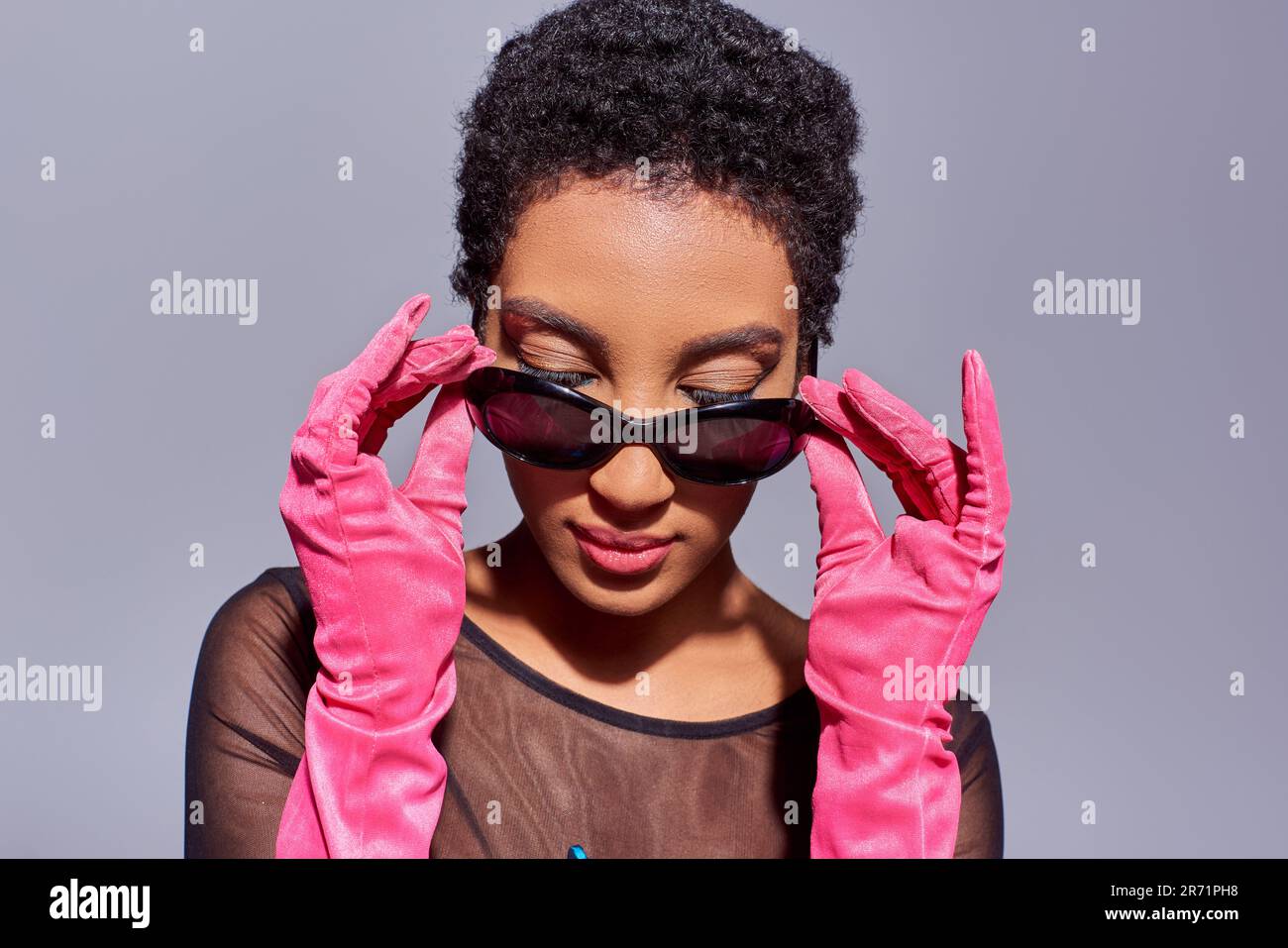 Giovane e sicura donna afroamericana che tocca gli occhiali da sole e indossa guanti rosa mentre si posa e si alza isolato sul grigio, moderna generazione z Foto Stock