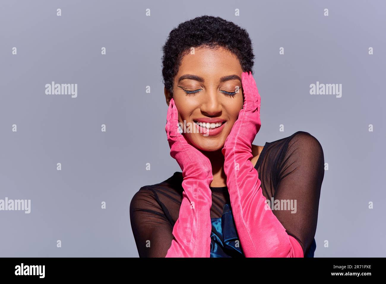 Gioiosa donna afroamericana con un trucco audace e guanti rosa che chiudono gli occhi e toccano il viso mentre si è in piedi isolato sul grigio, moderna generazione z fas Foto Stock