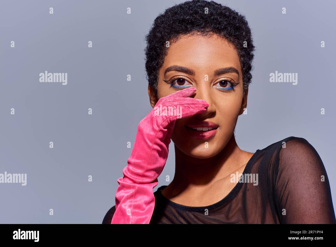 Ritratto di donna afroamericana alla moda con un trucco audace e un guanto rosa che tocca il viso e sorridente isolato sulla moda grigia e moderna della generazione z Foto Stock