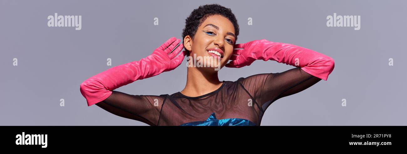 Donna afroamericana sorridente e alla moda con un trucco audace indossando guanti rosa e posando mentre si è in piedi isolato sulla moda grigia e moderna di generazione z Foto Stock