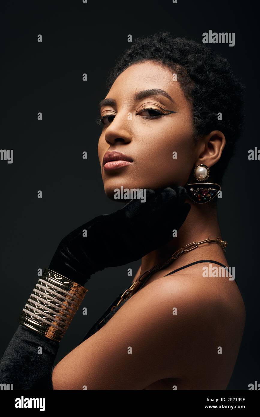 Ritratto di sicura ed elegante giovane afroamericana donna con trucco sera,  guanto e accessori dorati guardando la macchina fotografica isolato su nero  Foto stock - Alamy