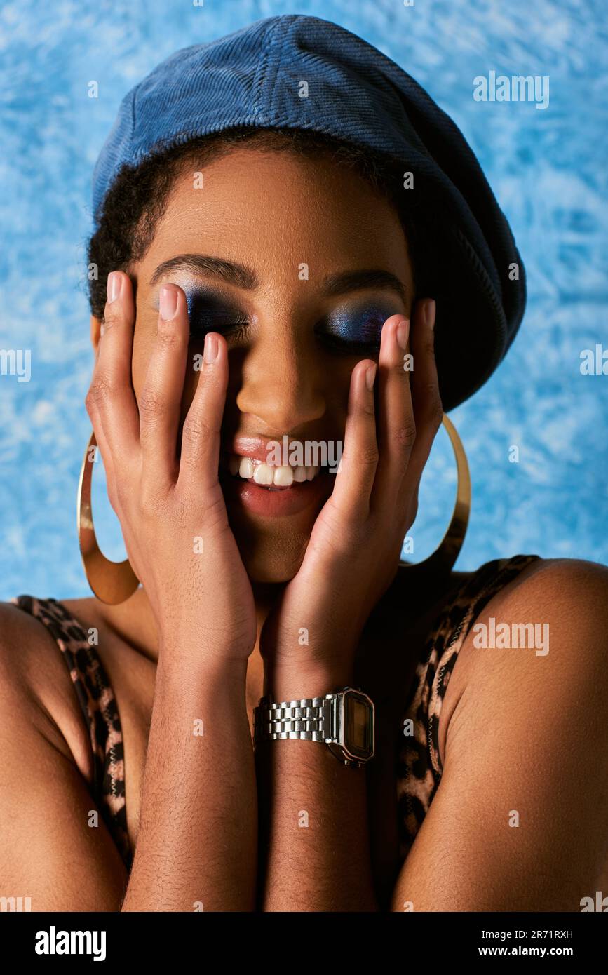 Ritratto di donna afro-americana positiva con trucco vivace e beretta toccare guance e chiudere gli occhi su sfondo blu textured, elegante denim a. Foto Stock
