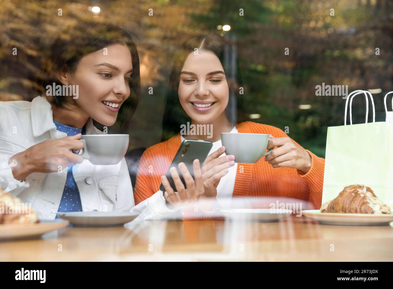 Promozione speciale. Donne giovani felici con tazze di bevande utilizzando smartphone al tavolo in caffè, vista dall'esterno Foto Stock Promozione speciale. Donne giovani felici con tazze di bevande utilizzando smartphone al tavolo in caffè, vista dall'esterno Foto Stock