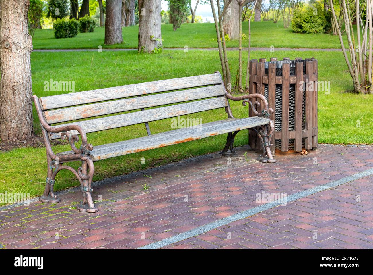 Una vecchia panca con tavole di legno intemperie e una base in bronzo metallico su un'area pavimentata sullo sfondo di un parco cittadino. Foto Stock