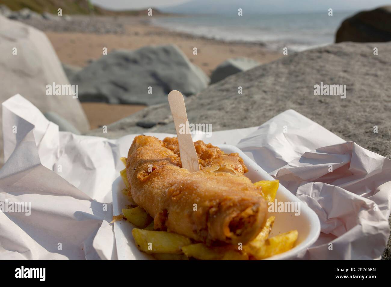Pesce e patatine, merluzzo in pastella, in un vassoio avvolto in carta con una forchetta di legno su una spiaggia. Cucina tradizionale britannica Foto Stock