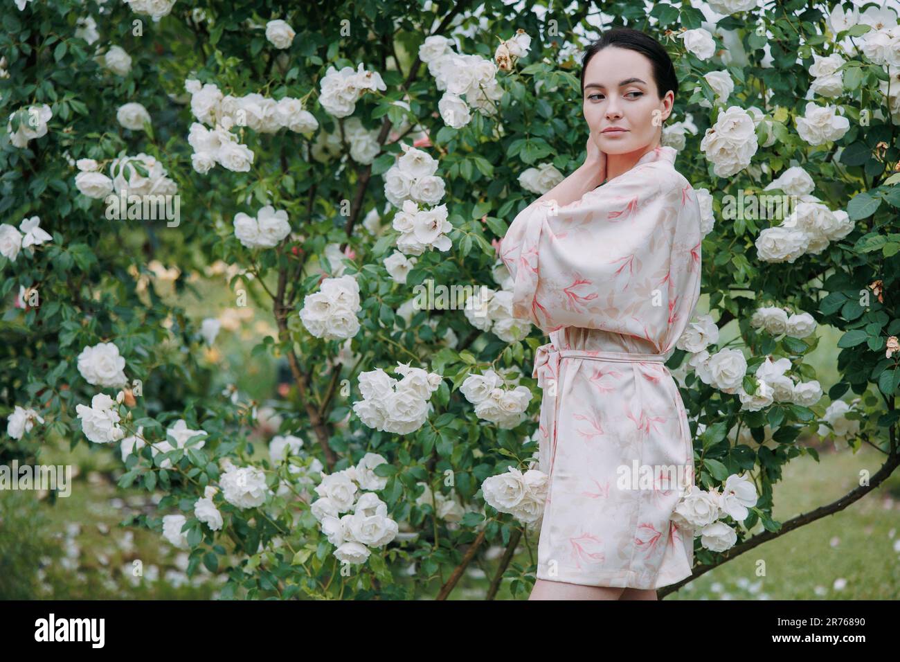 Giovane bruna donna in kimono vestito godendosi da arrampicata bianca Iceberg rosa in giardino. Foto Stock