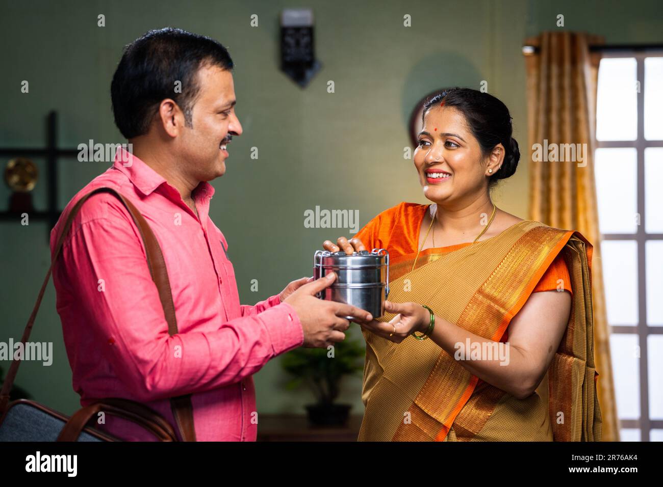 Donna indiana felice di mezza età che dà pranzo scatola a marito prima di andare in ufficio a casa - concetto di famiglia legame, cura e affettuoso. Foto Stock