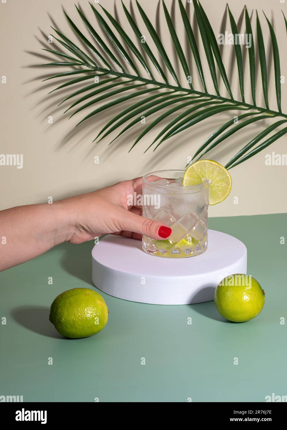 Anonimo femmina con smalto unghie rosso mettere vetro con cocktail drink con ghiaccio e fetta di agrumi in bianco mostra sul tavolo con limoni vicino al verde Foto Stock