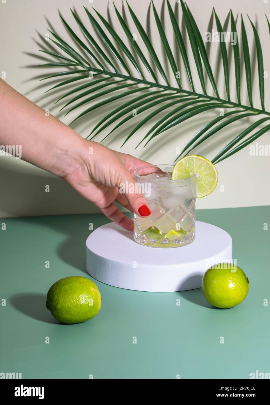 Anonimo femmina con smalto unghie rosso mettere vetro con cocktail drink con ghiaccio e fetta di agrumi in bianco mostra sul tavolo con limoni vicino alle foglie Foto Stock