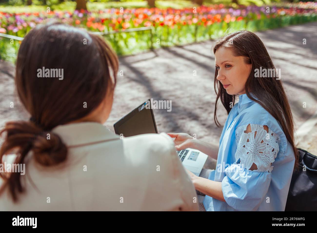 Due giovani donne d'affari che lavorano nel parco primaverile utilizzando un computer portatile e un libro. Lavoro a distanza durante la pausa. Amici che discutono delle attività. Partner del team meetin Foto Stock