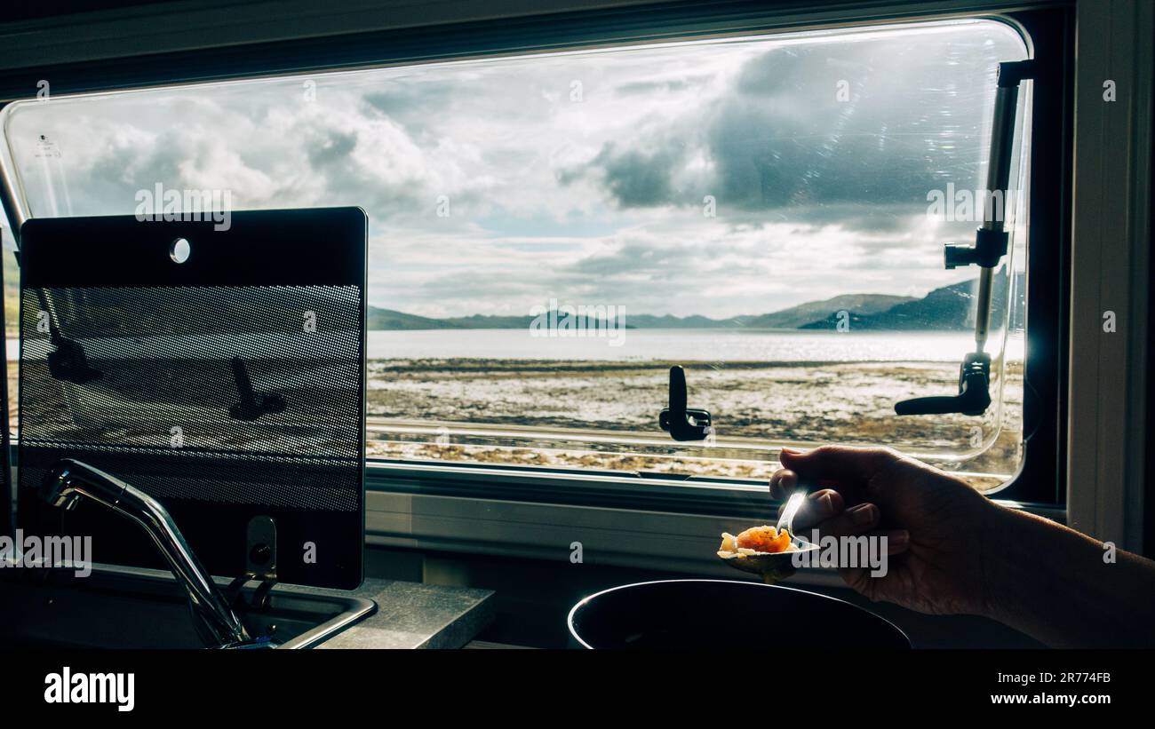 Persona che mangia la cena in un campervan che guarda fuori attraverso la finestra sopra un lago soleggiato nelle Highlands scozzesi su un campo selvaggio in Argyll. Scozia Foto Stock