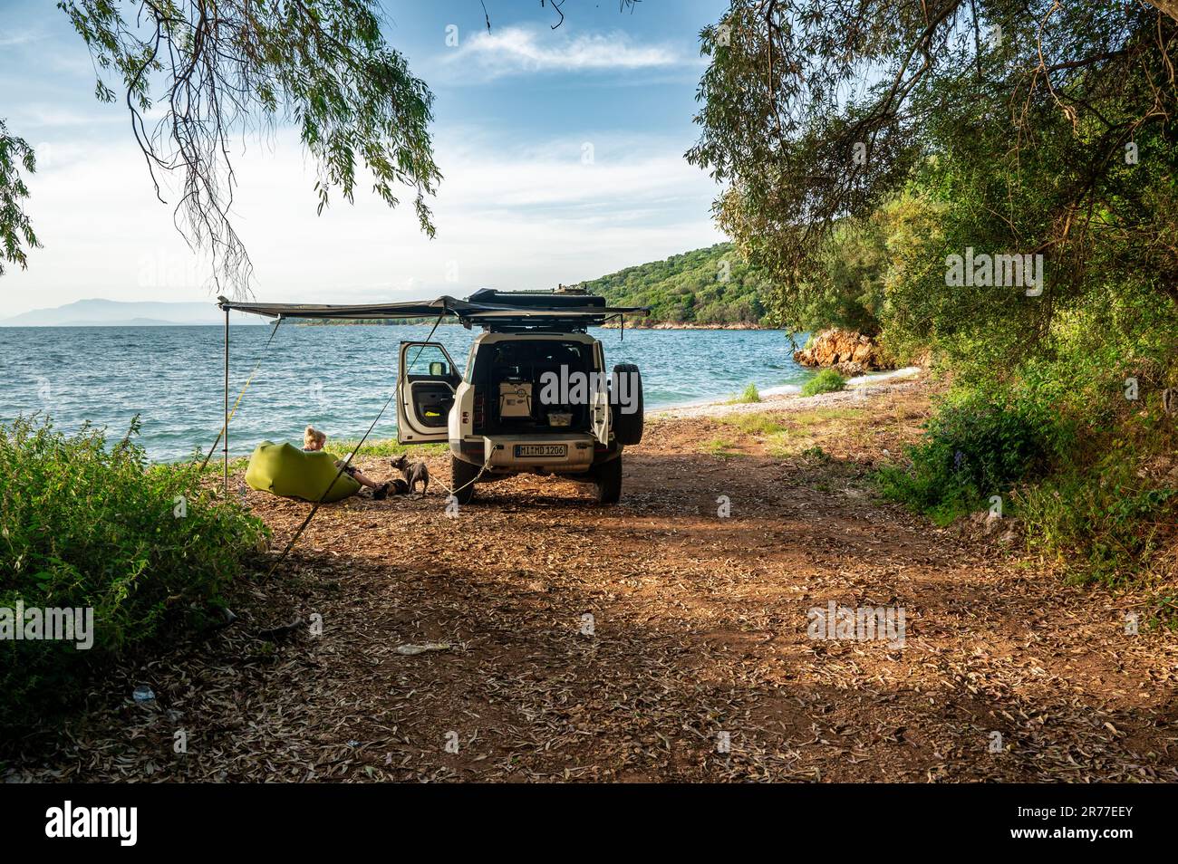 Campeggio per auto fuoristrada su una piccola spiaggia con limpide acque verdeggianti sulla costa del Peloponneso con donne e cani seduti davanti al veicolo Foto Stock