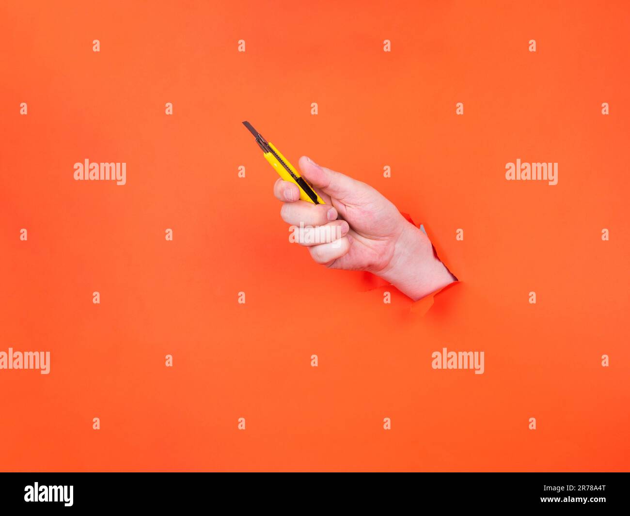 Una mano di un uomo tiene un taglierino giallo attraverso carta arancione strappata. Mano con coltello affilato. Foto Stock