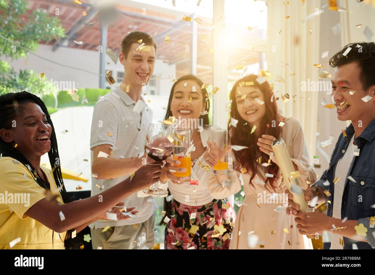 Gruppo di giovani amici interculturali eccitati che si aggettano con vino rosso in wineglass e succo d'arancia mentre si gode la festa di casa con i confetti Foto Stock