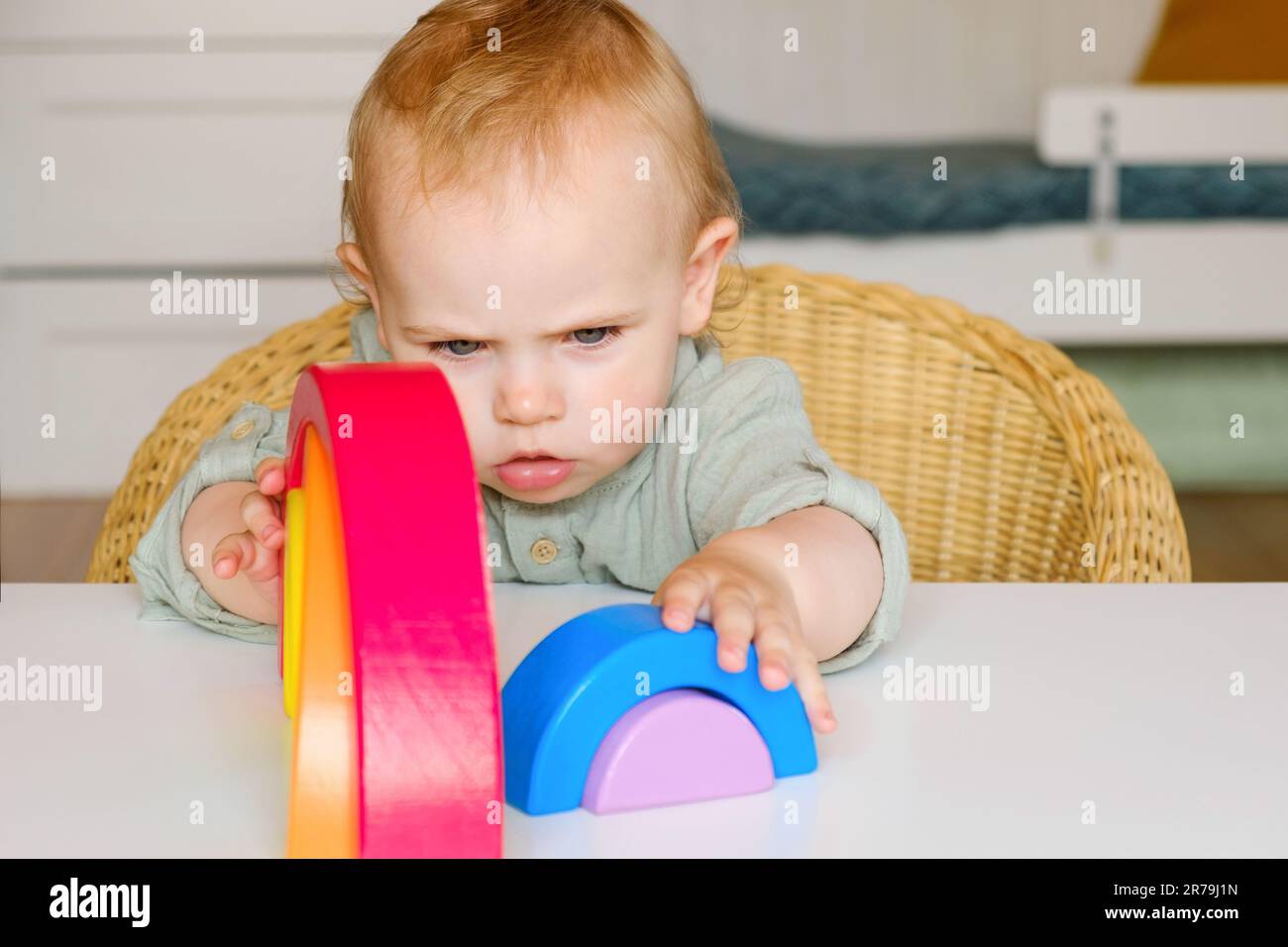 Bambino piccolo in vestiti fatti di tessuto naturale gioca con i giocattoli di legno colorati arcobaleno al tavolo bianco. Camera accogliente in stile scandinavo. Foto Stock