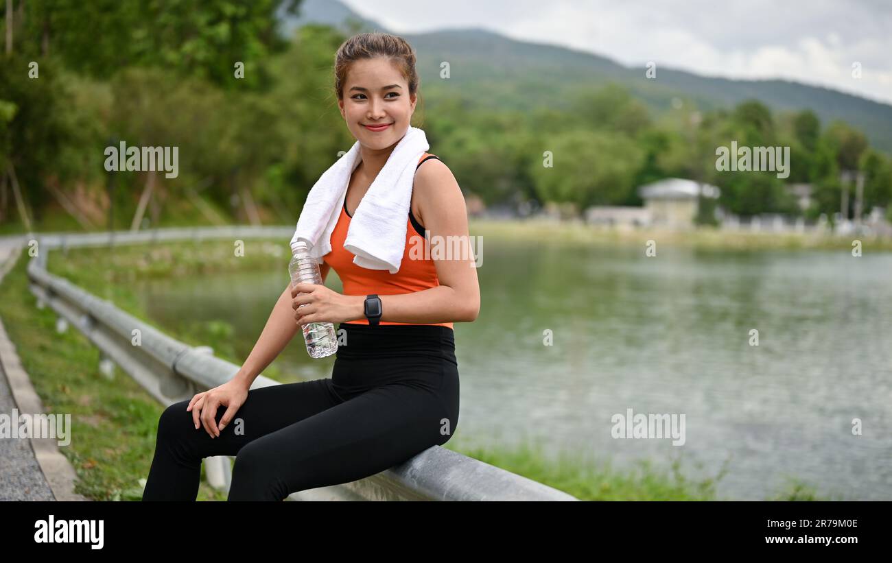 Una bella e felice donna asiatica millenaria in abbigliamento sportivo si riposa dopo una lunga corsa nel parco pubblico. allenamento, cardio, esercizio, tempo libero, wellbei Foto Stock