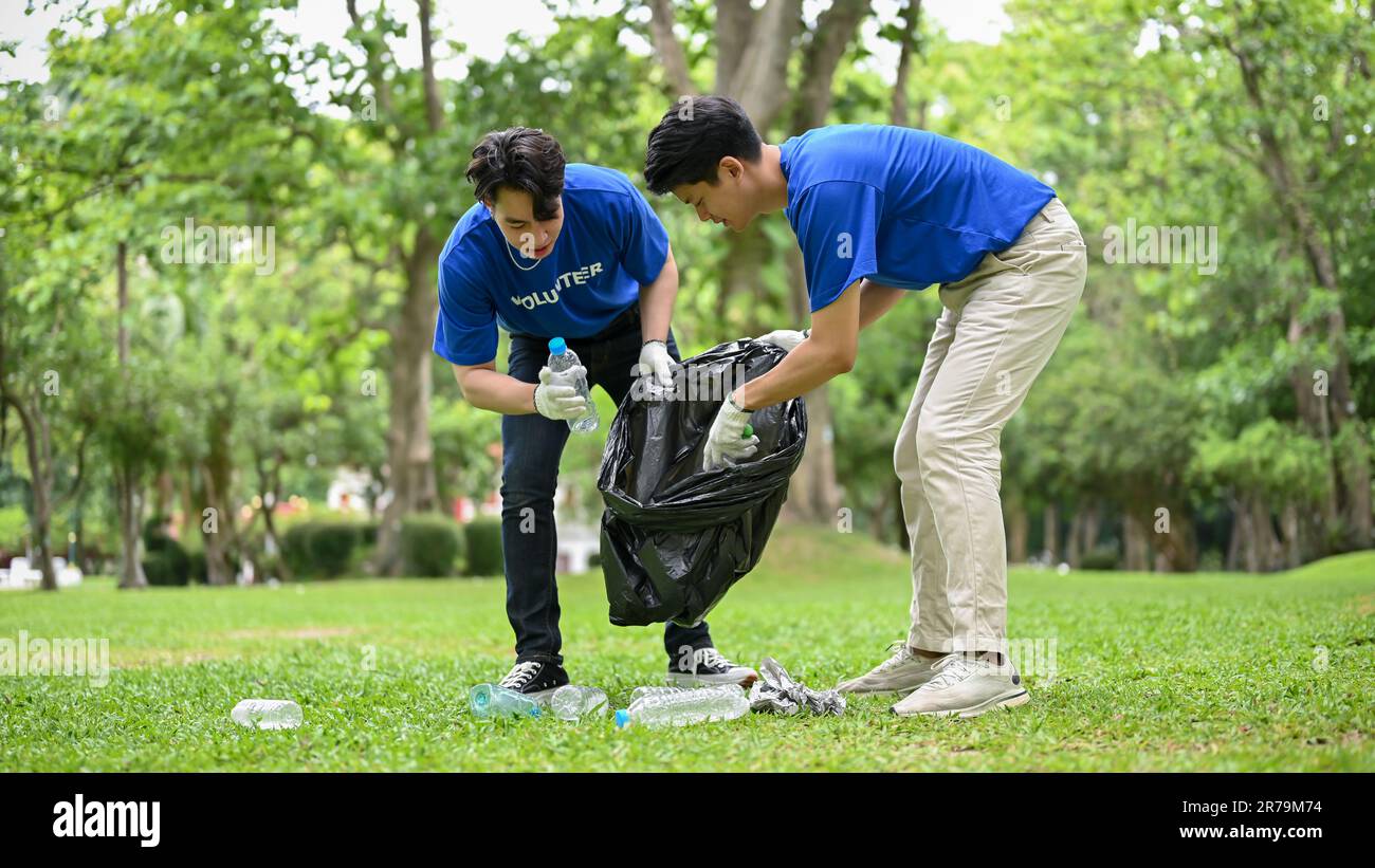 Due giovani uomini asiatici felici con sacco spazzatura aiutare a vicenda pulire la zona in un parco pubblico. Beneficenza comunitaria, volontariato, riciclaggio, csr pr Foto Stock