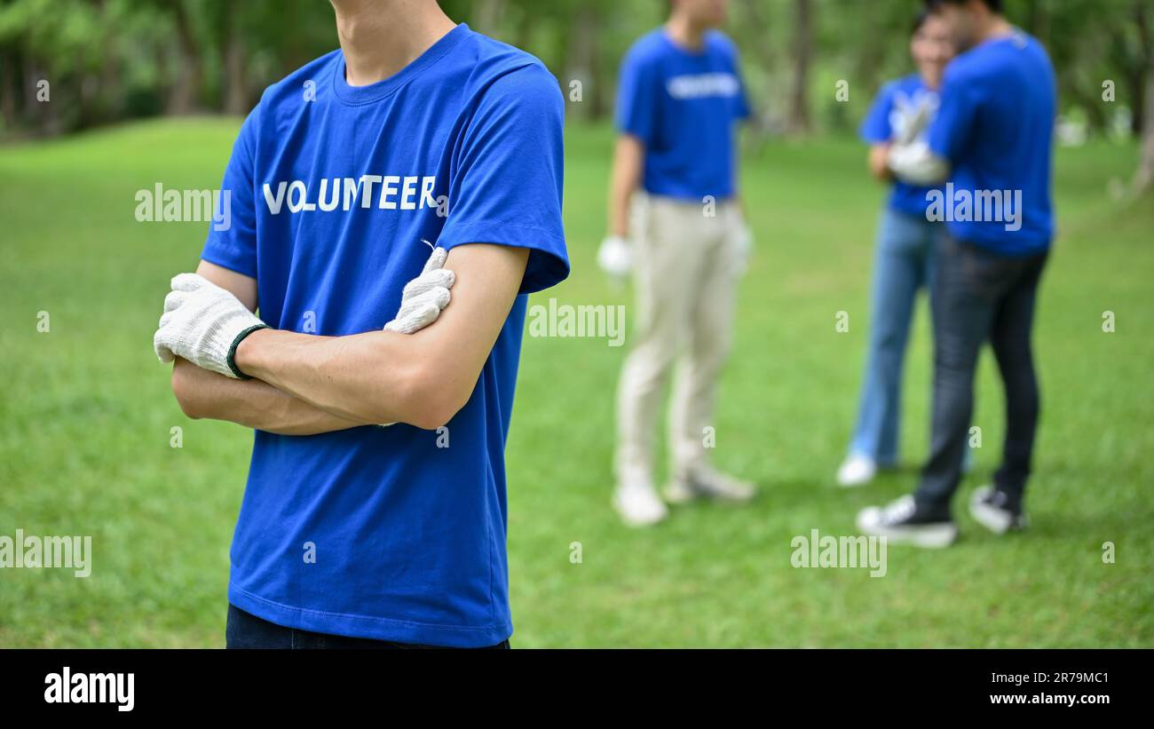 Immagine ritagliata di un volontario maschile asiatico in uniforme si trova in un parco pubblico con le braccia incrociate. Volontariato, carità comunitaria, ambiente, donat Foto Stock