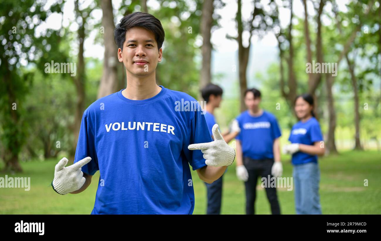 Un giovane volontario asiatico in uniforme, felice e bello, si trova in un parco pubblico, partecipando al lavoro volontario con i suoi amici per pulire un pubblico Foto Stock