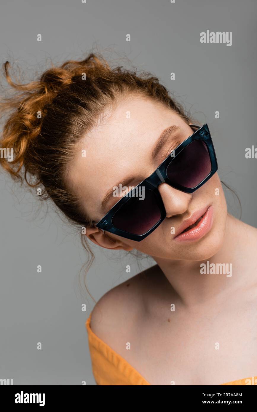 Ritratto di donna alla moda con capelli rossi e giovani con trucco naturale indossando occhiali da sole in piedi isolati su sfondo grigio, alla moda sole prote Foto Stock