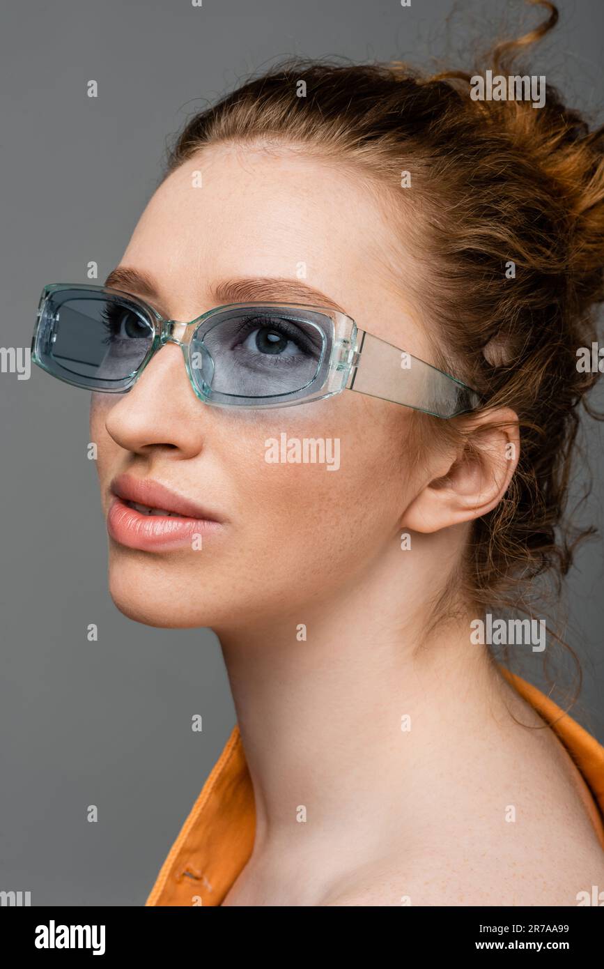 Ritratto di giovane donna alla moda arrossata con lenticchi e trucco naturale indossando occhiali da sole blu e guardando lontano isolato su sfondo grigio, tre Foto Stock