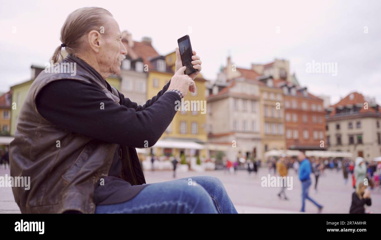 Senior siede sui gradini e cattura le attrazioni turistiche utilizzando uno smartphone nel centro storico di una vecchia città europea. Piazza del Palazzo, Città Vecchia di Varsavia Foto Stock