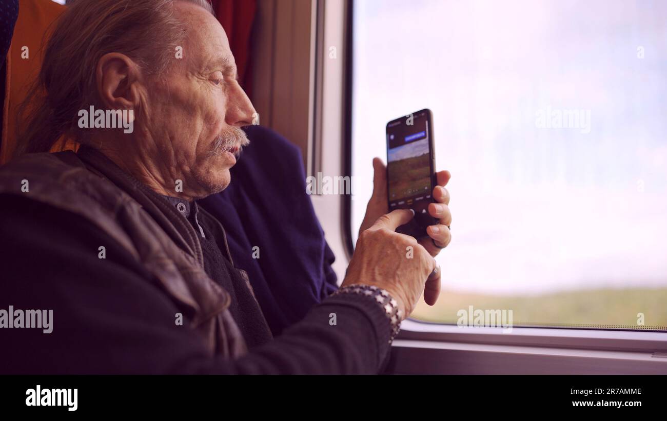Uomo anziano che viaggia in treno e scatta foto del paesaggio attraverso la finestra utilizzando uno smartphone Foto Stock