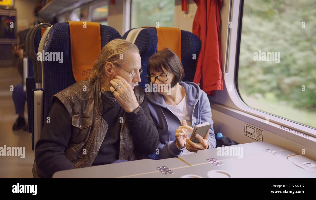La coppia anziana viaggia in treno, la signora tiene in mano un telefono cellulare, entrambi guardano lo smartphone e parlano tra loro Foto Stock