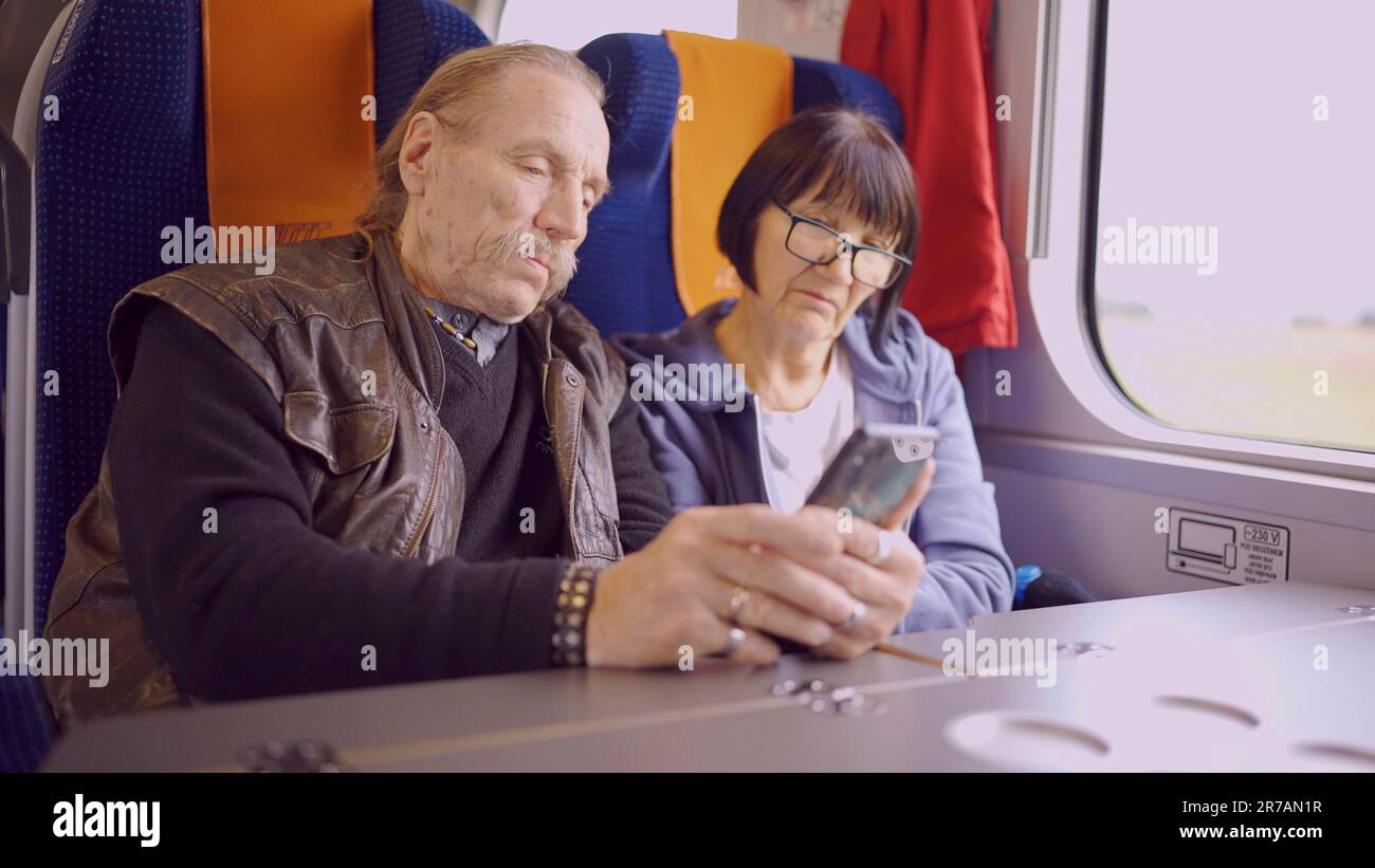 La coppia anziana viaggia in treno, l'anziano tiene in mano un telefono cellulare, entrambi guardano lo smartphone e parlano l'uno con l'altro Foto Stock