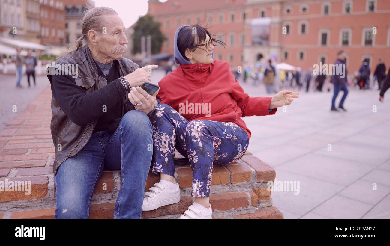 Una coppia di turisti anziani è seduta e parla nel centro storico di una vecchia città europea, senior ha uno smartphone in mano. Piazza del Palazzo, Foto Stock