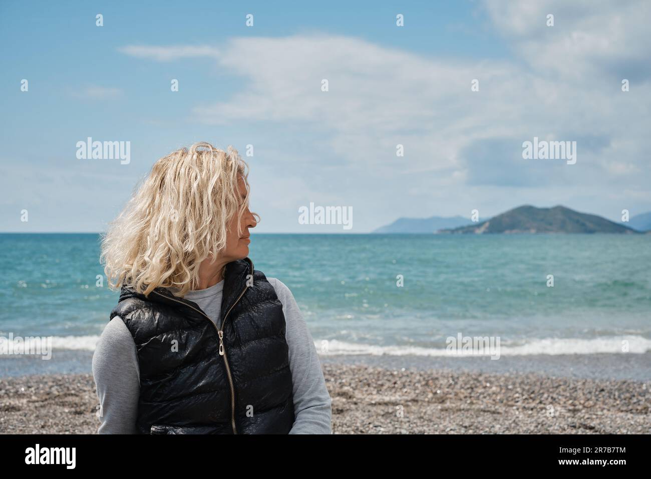 donna europea bionda adulta seduta da sola su una spiaggia di ciottoli con mare blu e cielo sullo sfondo, guardando lontano. Persone in vacanza e in vacanza, lif Foto Stock
