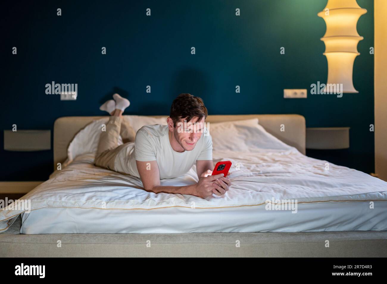 Uomo che legge le informazioni in smartphone sdraiato a letto a casa in camera da letto moderna di sera. Foto Stock