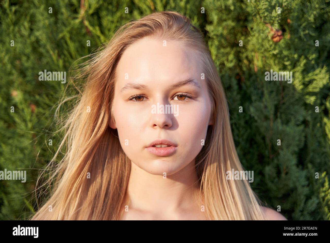 Una giovane donna sta in un giardino, le braccia incrociate e la testa leggermente inclinata, guardando verso la telecamera con un sorriso dolce Foto Stock