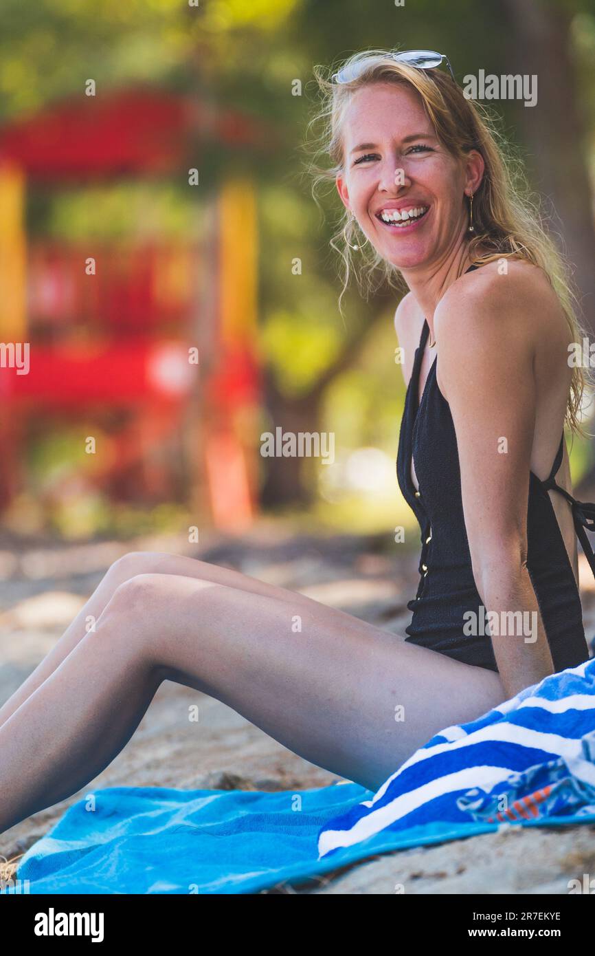 Donna felice sorridente alla macchina fotografica sulla spiaggia tropicale seduta in asciugamano da bagno Foto Stock