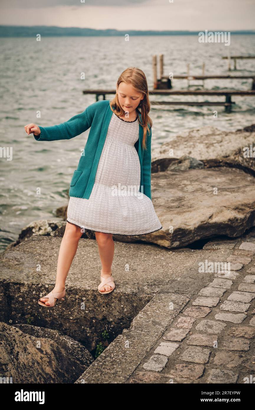 Ritratto all'aperto di adorabile bambina che gioca accanto al lago, indossando un abito bianco e una giacca verde cardigan Foto Stock