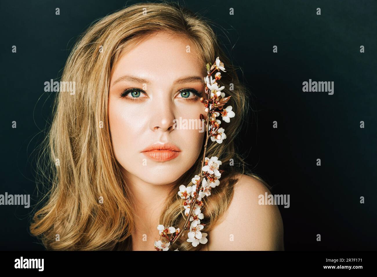 Primo piano ritratto di bella giovane donna con capelli biondi e make up professionale, posa su sfondo nero, tenendo fiorito il ramo primaverile di pisello Foto Stock