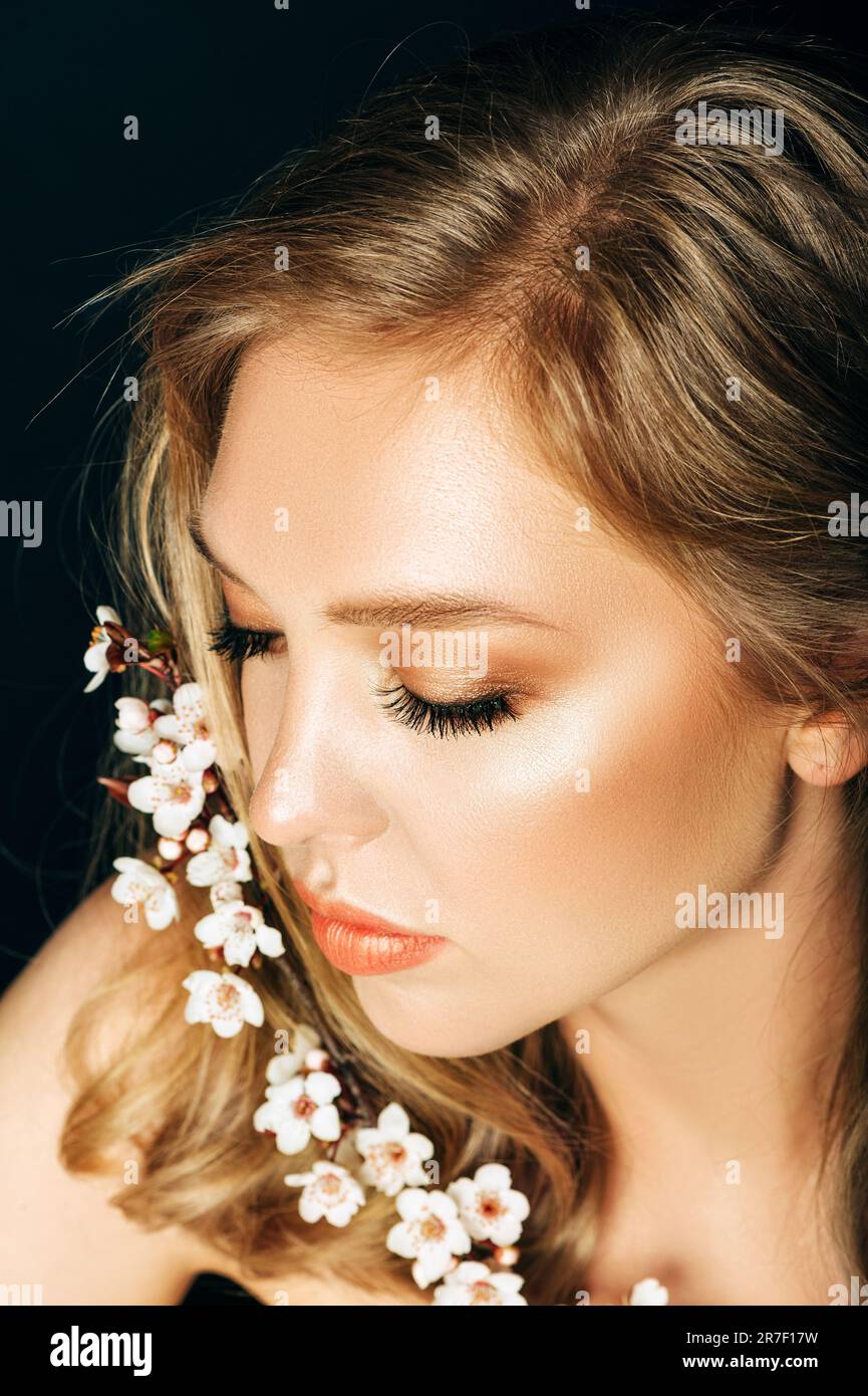 Primo piano ritratto di bella giovane donna con capelli biondi e make up professionale, posa su sfondo nero, tenendo fiorito il ramo primaverile di pisello Foto Stock