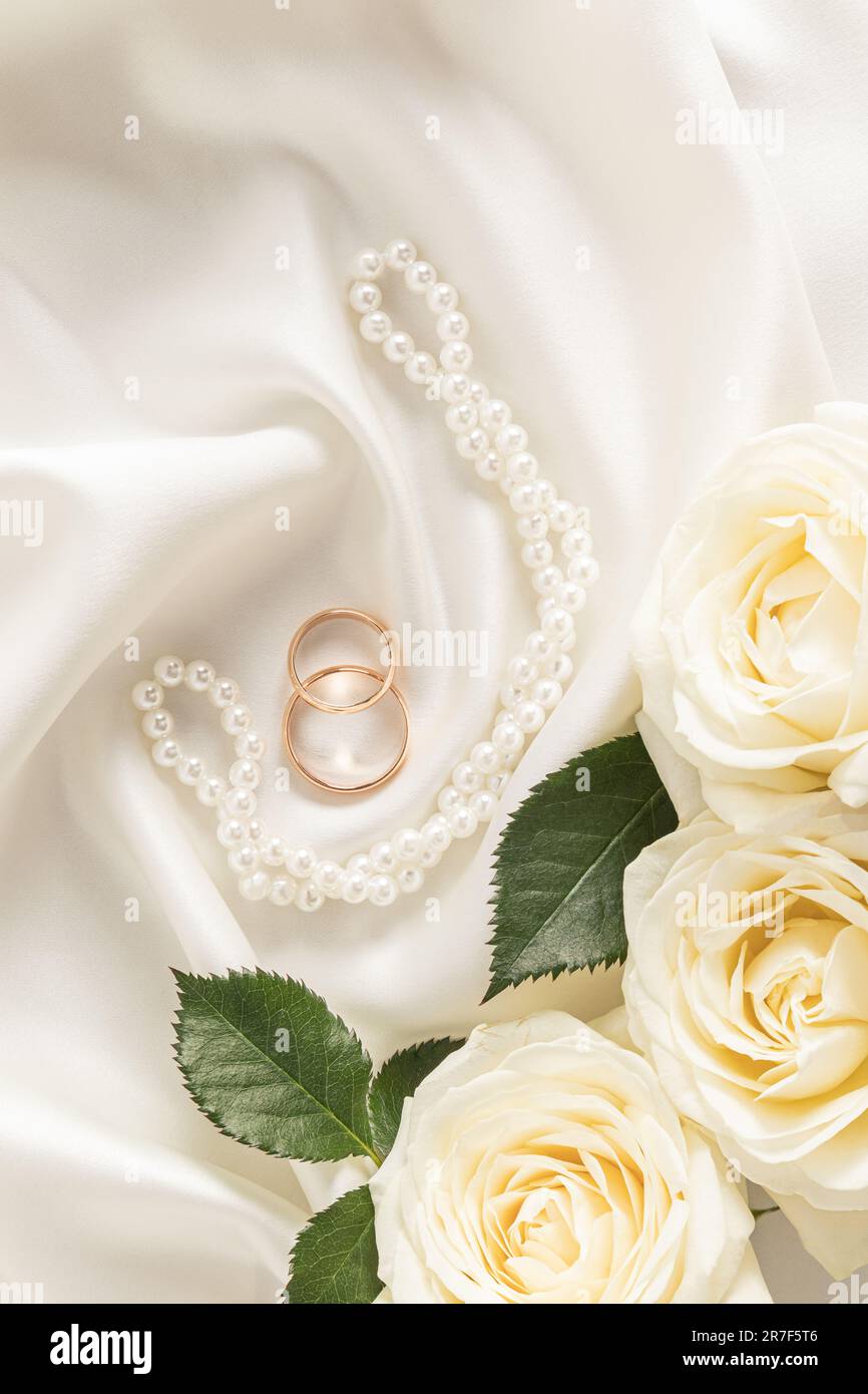 Due anelli d'aggancio dorati su fondo avorio satinato con perle e rose bianche. Vista verticale dall'alto. layout per la progettazione Foto Stock