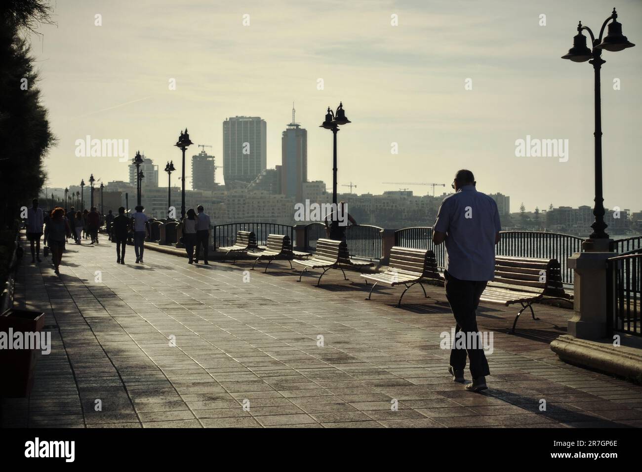 Persone che camminano alla luce del sole dietro sul lungomare di Sliema, sullo sfondo lo skyline di St. Julian's, Malta Foto Stock