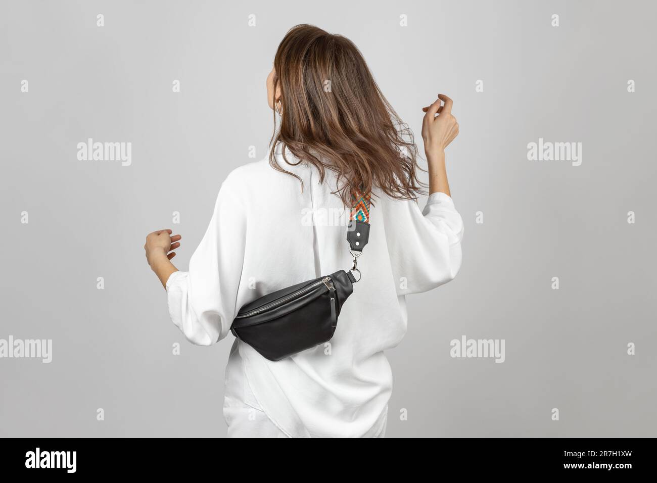 Donna bianca che indossa jeans bianchi, camicia e tracolla Borsa da cintura in pelle nera su sfondo grigio Foto Stock