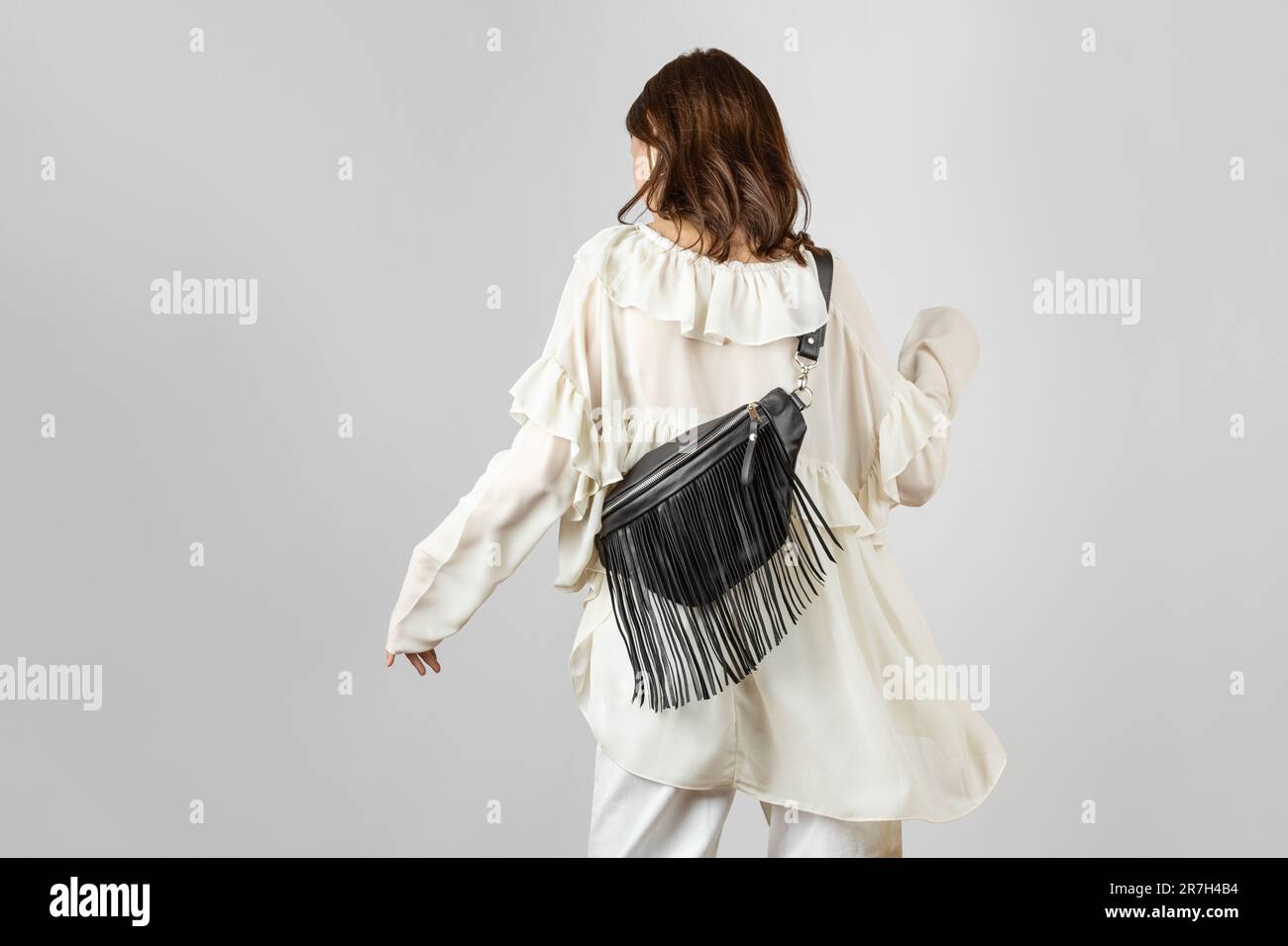 Donna bianca da dietro che indossa abiti bianchi e tracolla Borsa da cintura in pelle nera con frangia decorativa in stile Boho su sfondo grigio Foto Stock