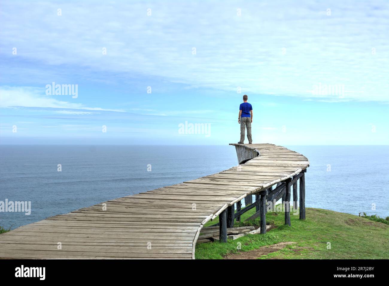 Un uomo si trova sul bordo di una lunga passerella in legno (il "Dock of Souls" di Chiloé) e guarda l'oceano all'orizzonte in una giornata limpida Foto Stock
