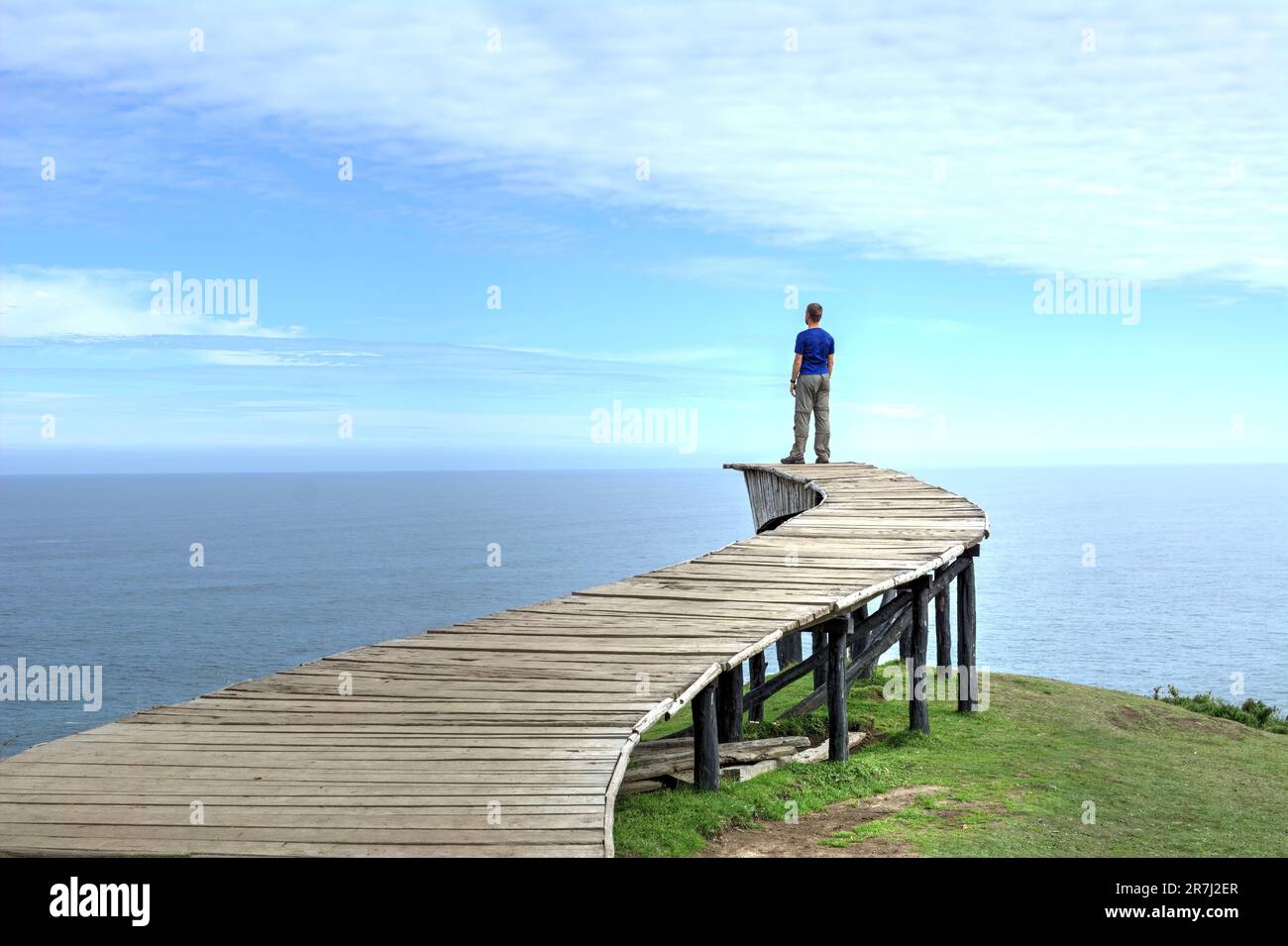 Un uomo si trova sul bordo di una lunga passerella in legno (il "Dock of Souls" di Chiloé) e guarda l'oceano all'orizzonte in una giornata limpida Foto Stock