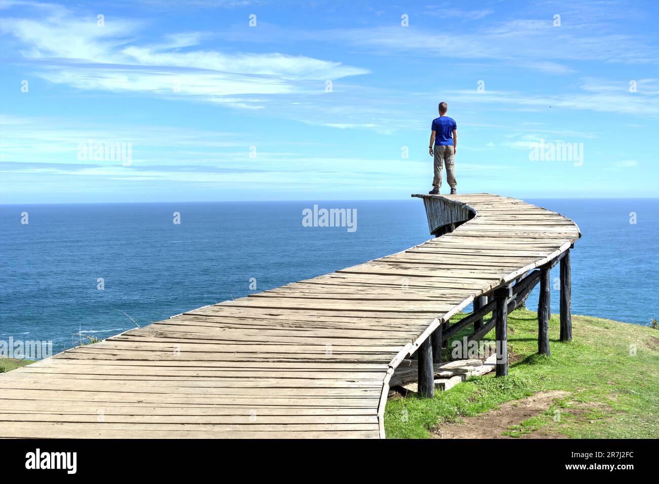 Un uomo si trova sul bordo di una lunga passerella in legno (il "Dock of Souls" di Chiloé) e guarda l'oceano all'orizzonte in una giornata limpida Foto Stock