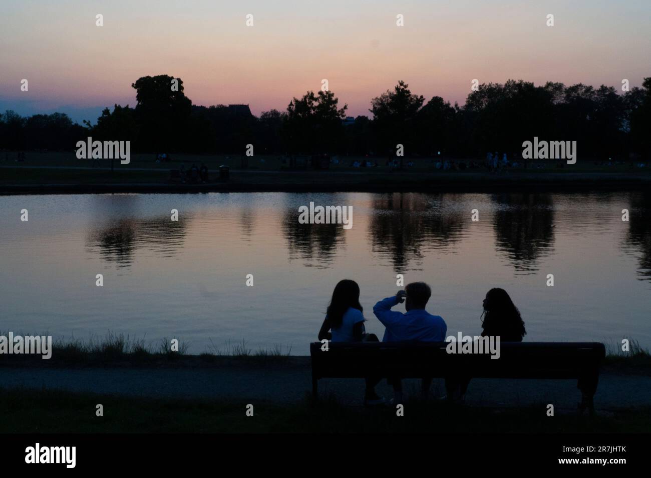 UK Weather, Londra 15 giugno 2023: Durante e prolungato periodo di caldo clima secco con temperature giornaliere elevate di 27 centigradi, tre persone si siedono su una panchina chiacchierando al crepuscolo, mentre i colori sbiaditi del tramonto si riflettono in Long Pond, Clapham Common. Credit: Anna Watson/Alamy Live News Foto Stock