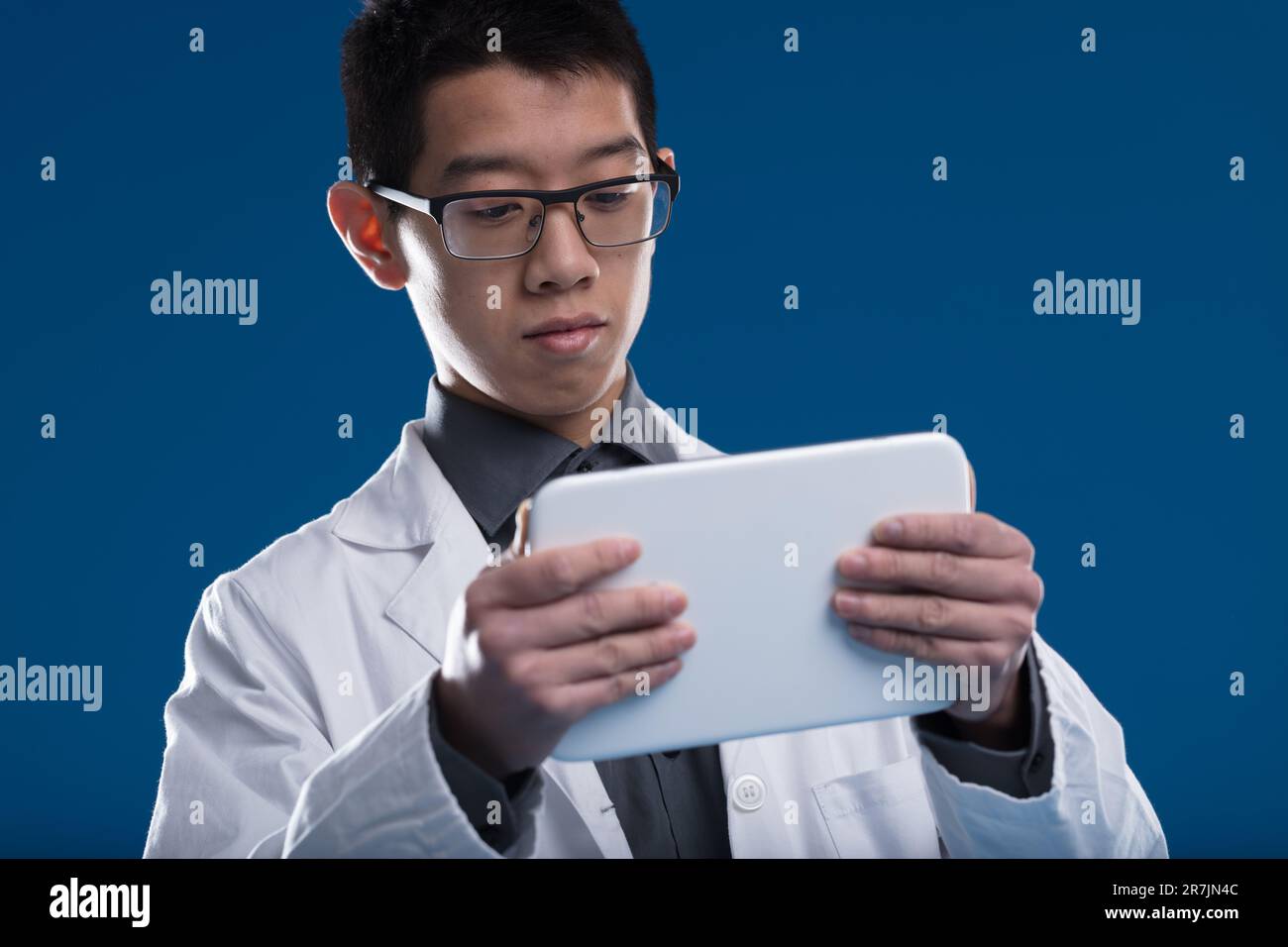 Lo scienziato asiatico perusa gli aggiornamenti e i dati sul suo tablet. Un ricercatore di etnia cinese, posto su uno sfondo blu. Indossare occhiali, intrigati dalla scr Foto Stock